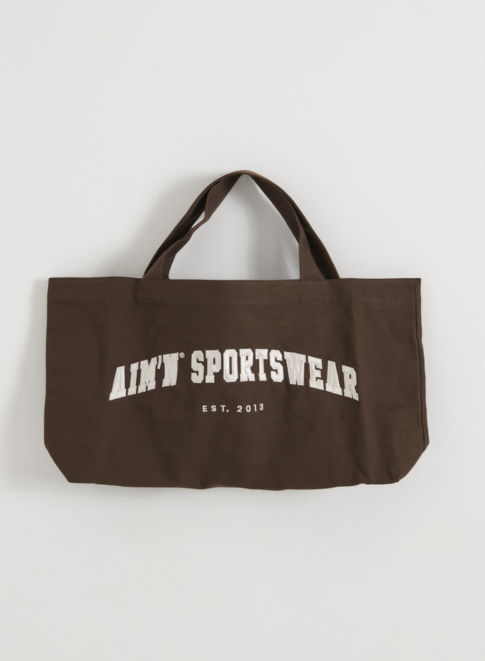 Chocolate brown cotton varsity tote bag with white aim'n logo lettering, shown in a flatlay.