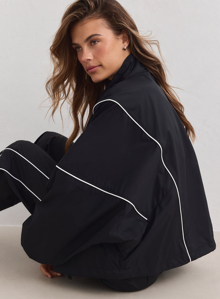 Black Balance Windstopper Tracksuit Jacket with white piping by aim'n, styled in a loose fit on a sitting model.