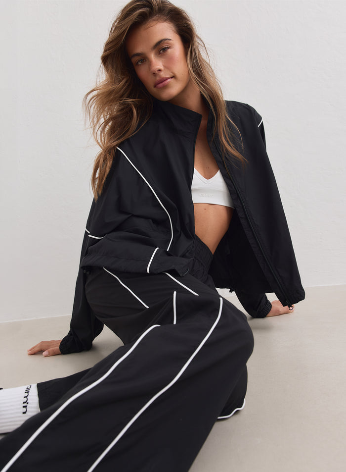 Black aim'n Balance Windstopper Tracksuit Jacket with white piping, worn by a model sitting down.