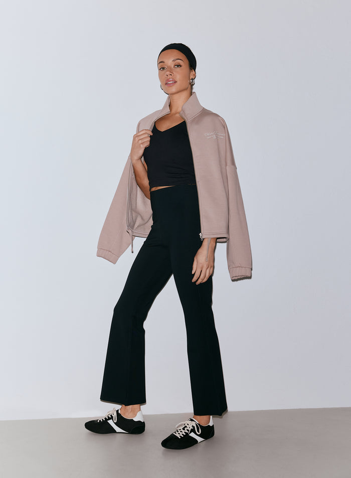 Woman wearing black Luxe Kick Flare Leggings, black crop top, and a light brown zip-up jacket draped over her shoulders, with a black headband and black and white sneakers. [36-170cm]