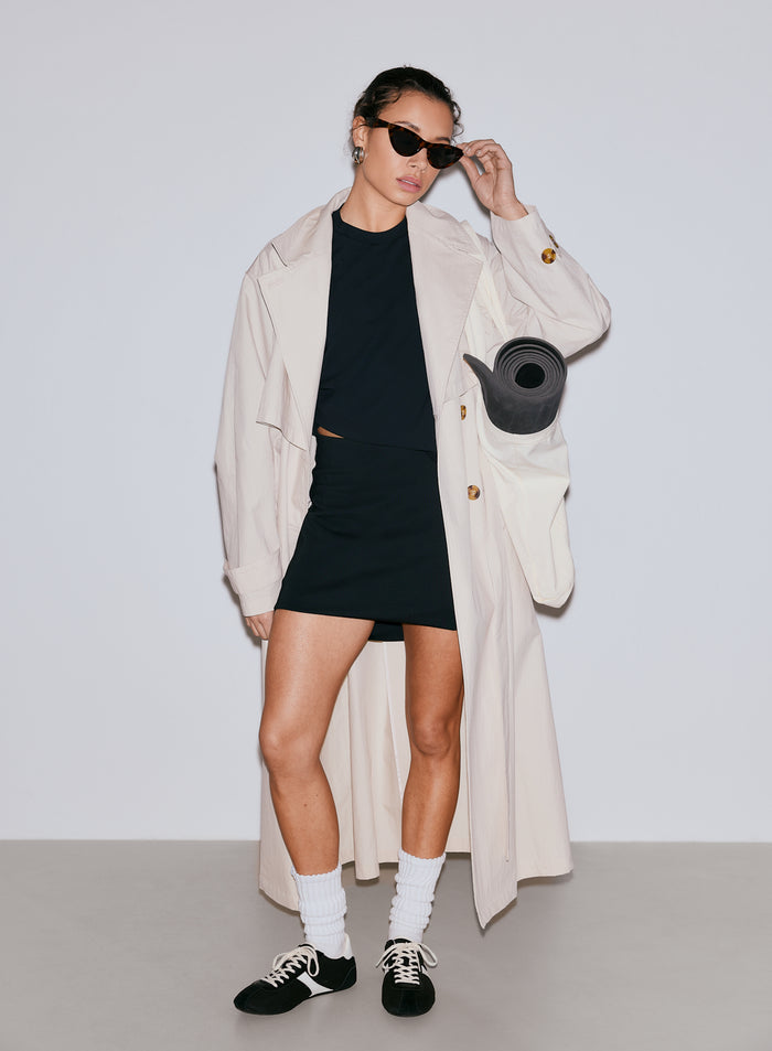 Woman in black Luxe Mini Skort, black top, and beige trench coat with a yoga mat [36-170CM]