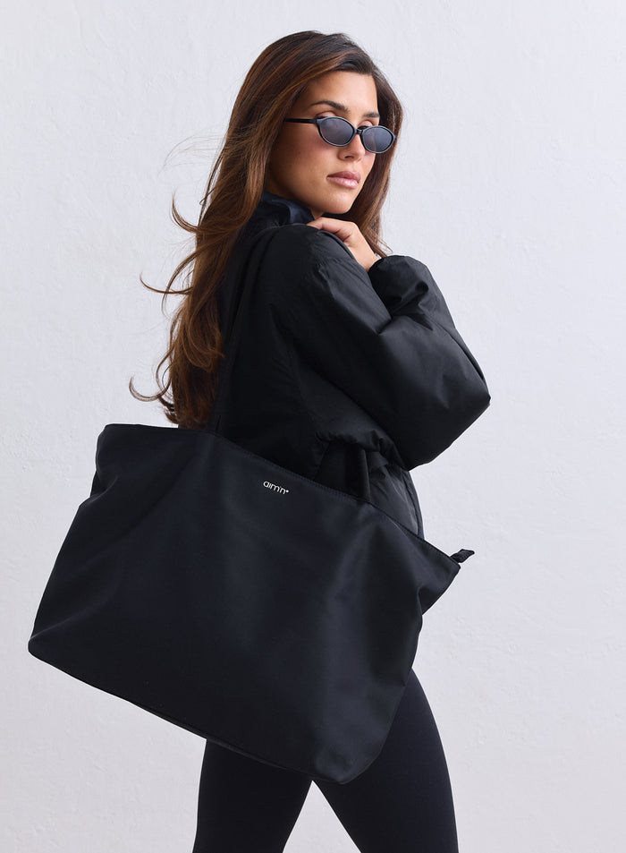 Large black nylon tote bag with a small aim'n logo carried by a woman wearing a black jacket.