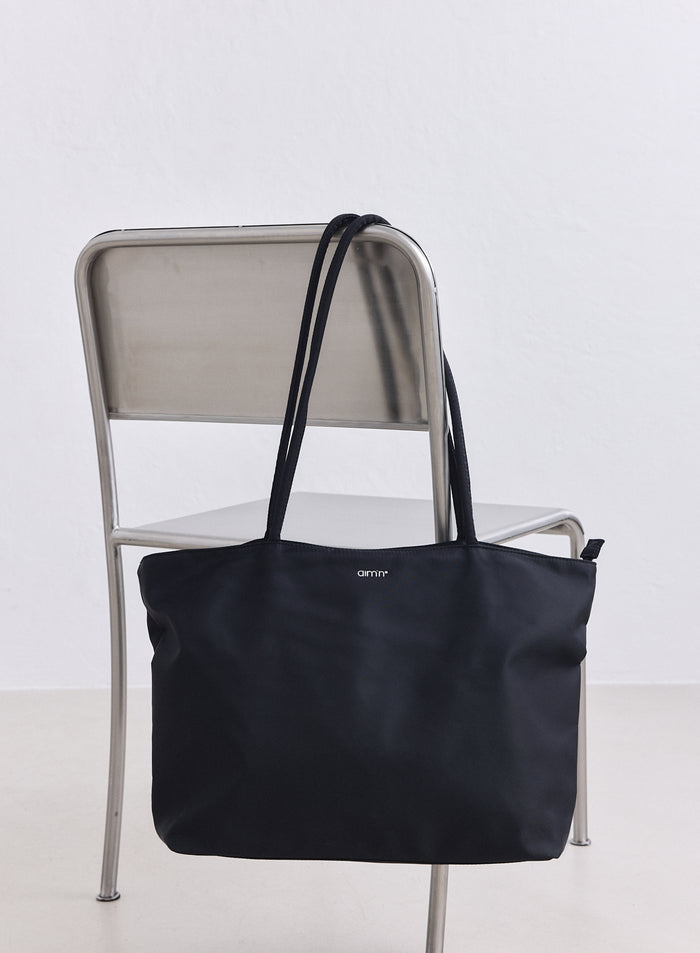Black nylon tote bag with white aim'n logo hanging on a minimalist metal chair.