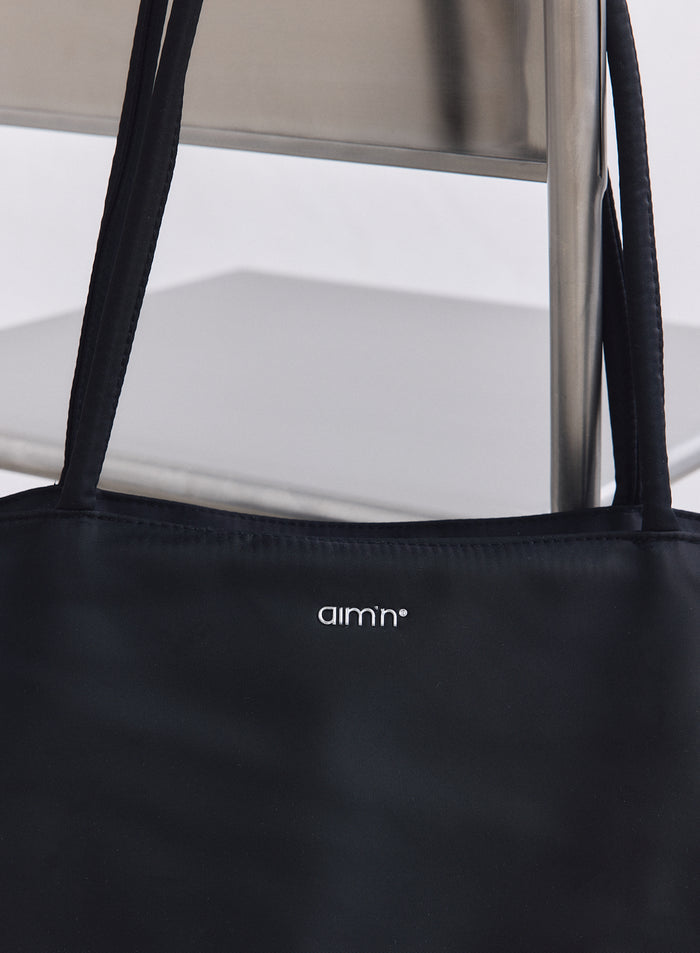 Minimalist black nylon tote bag from aim'n with silver logo and long straps on a metal chair.
