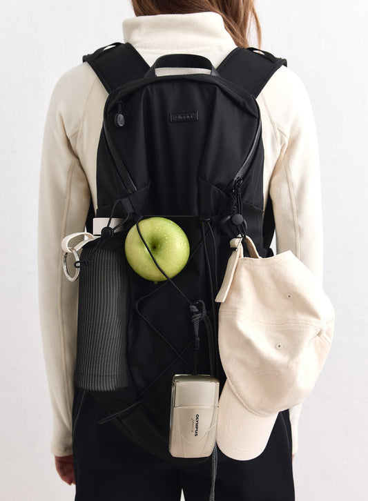 Black Outdoor Daypack by aim'n with a green apple, water bottle, cap, and camera attached to cords.