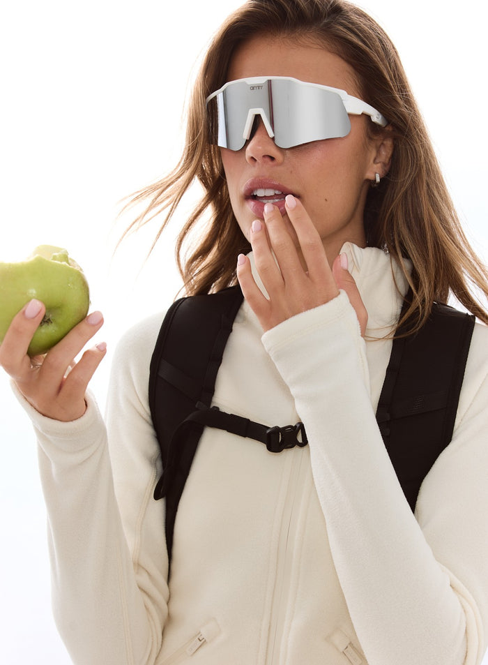 Black aim'n Outdoor Daypack worn by a woman in a white fleece jacket and white sunglasses holding a green apple.