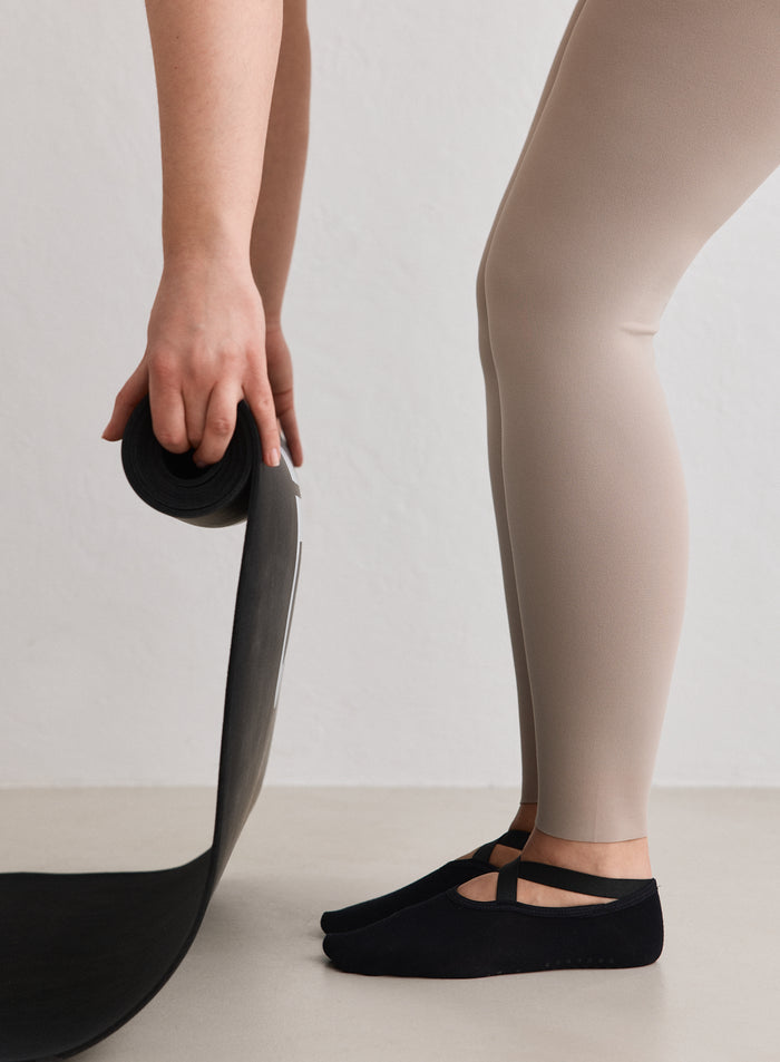 A person wearing black Pilates Socks from aim'n and beige leggings, holding a rolled-up black exercise mat.