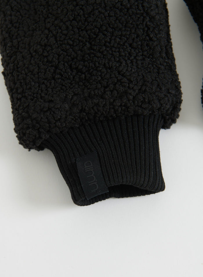 Close-up of a black pile mitten with a textured fleece surface, ribbed cuff and aim'n label