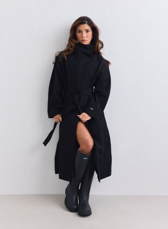 Long black waterproof coat by aim'n featuring a tie waist and paired with tall rubber boots. [S-165cm]