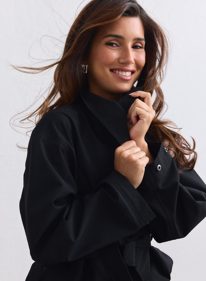 Smiling model wearing a black aim'n waterproof coat while holding up the collar. [S-165cm]