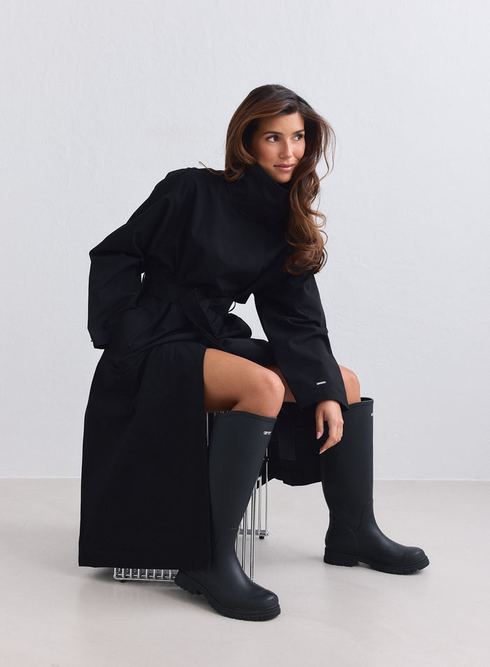 Long black waterproof coat from aim'n with a high collar, worn with rain boots. [S-165cm]
