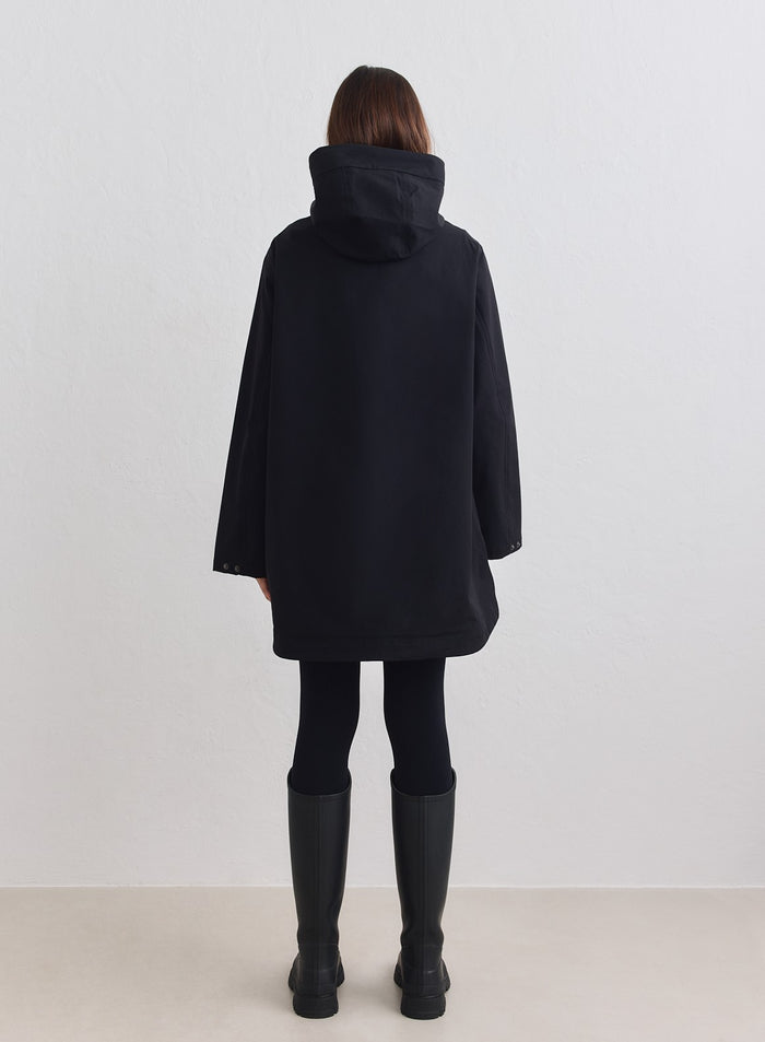 Black aim'n waterproof hood jacket with a long, minimalist fit, shown from the back with rain boots. [M-178cm]