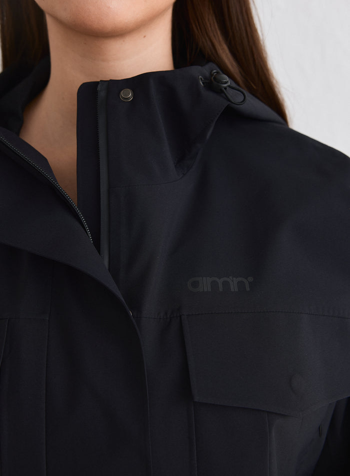 Close-up of the Black Waterproof Jacket showing the tonal reflective aim'n logo, snap buttons, and hood puller [M-174cm]