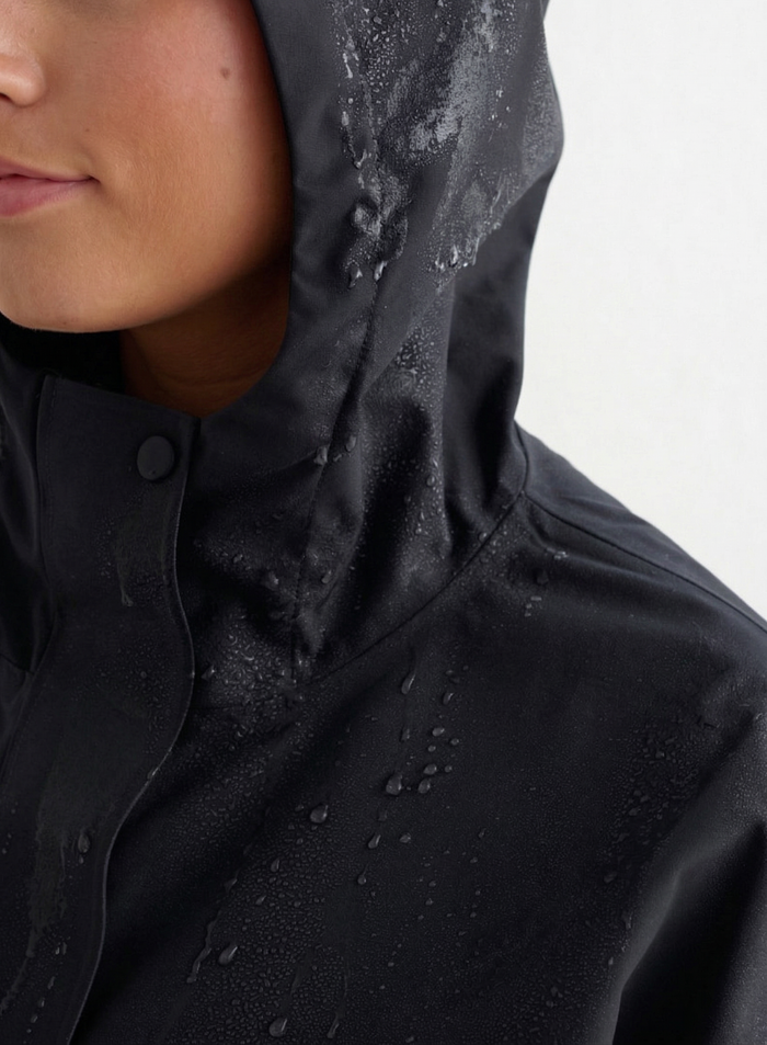 Close-up of a person wearing a black waterproof oversized anorak with water droplets on the hood and fabric