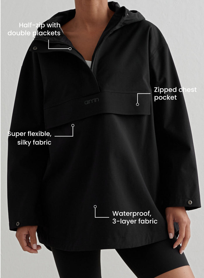 A close-up of a person wearing a black Waterproof Oversized Anorak from aim'n, featuring callouts highlighting functional details such as the half-zip with double plackets, zipped chest pocket, and waterproof 3-layer fabric.