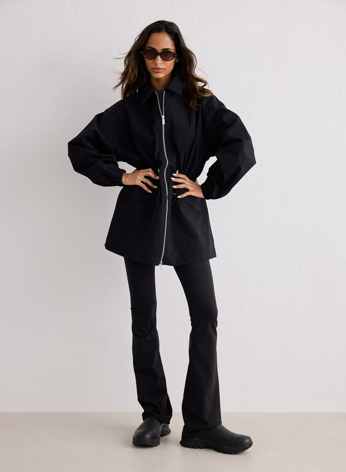 A female model wearing a black waterproof zip jacket with a collar, double zippers, and an adjustable cinched waist, paired with black flared trousers and chunky boots [S-170cm]