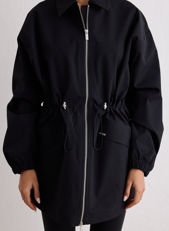 Cropped indoor studio shot of a model wearing the black Waterproof Zip Jacket, featuring a silver full zip extending from the bottom of the jacket to the collar, with the silver zip pull tab clearly visible. A drawstring waist with silver metal toggles accentuates the jacket, and the model's hand with light pink manicured nails rests at the bottom right of the image. [S-170cm]