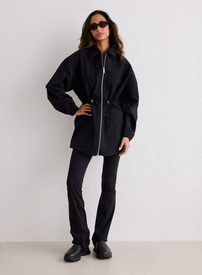 A woman posing in a black waterproof zip jacket with cinched waist, black pants, and sunglasses [S-170CM]