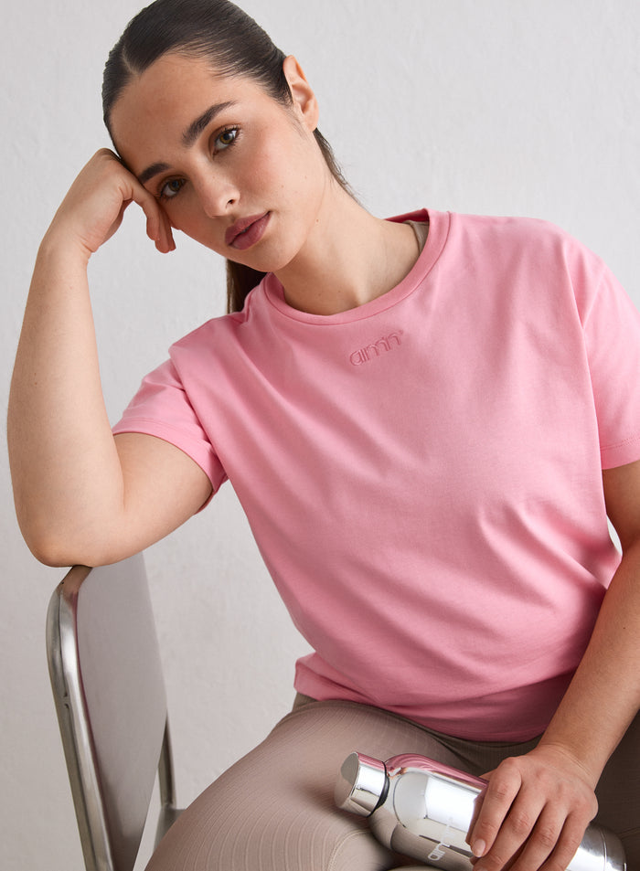 A woman wearing a Blush Boxy T-shirt with an embroidered aim'n logo sitting on a chair holding a silver water bottle [L-174cm]