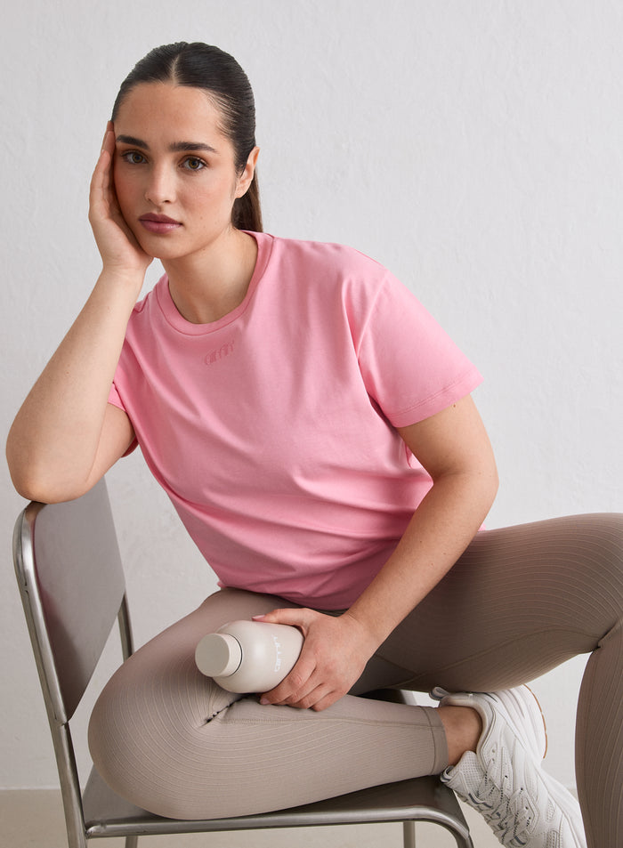 A woman sitting on a chair wearing a Blush Boxy T-shirt with an embroidered aim'n logo [L-174cm]