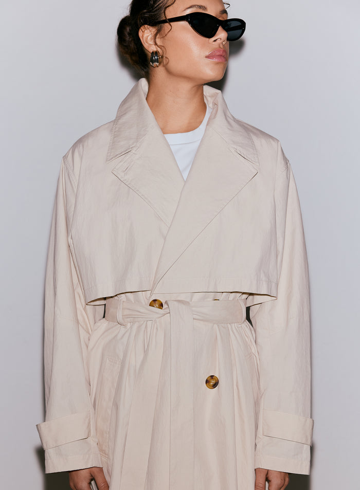 Close up of a woman wearing a café au lait aim'n Femme Trenchcoat, featuring a tied belt and large buttons, paired with black sunglasses. [M-170cm]