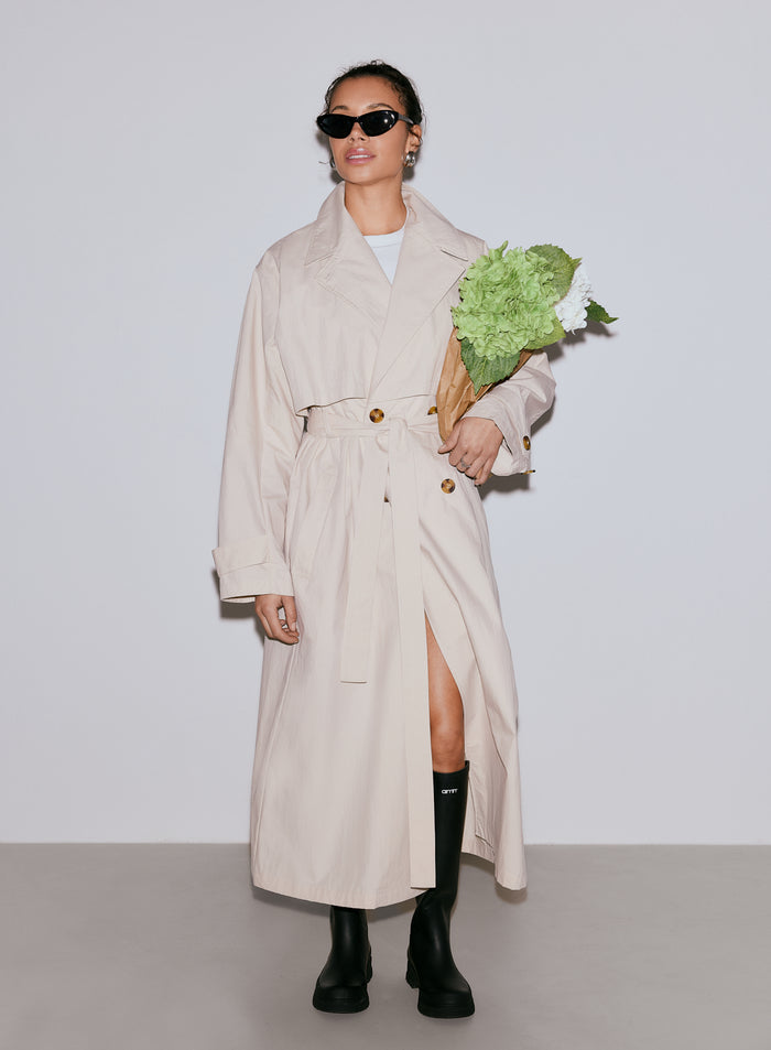 A woman wearing a long café au lait Femme Trenchcoat, black sunglasses, and black boots, holding a bouquet of green and white flowers [M-170cm]