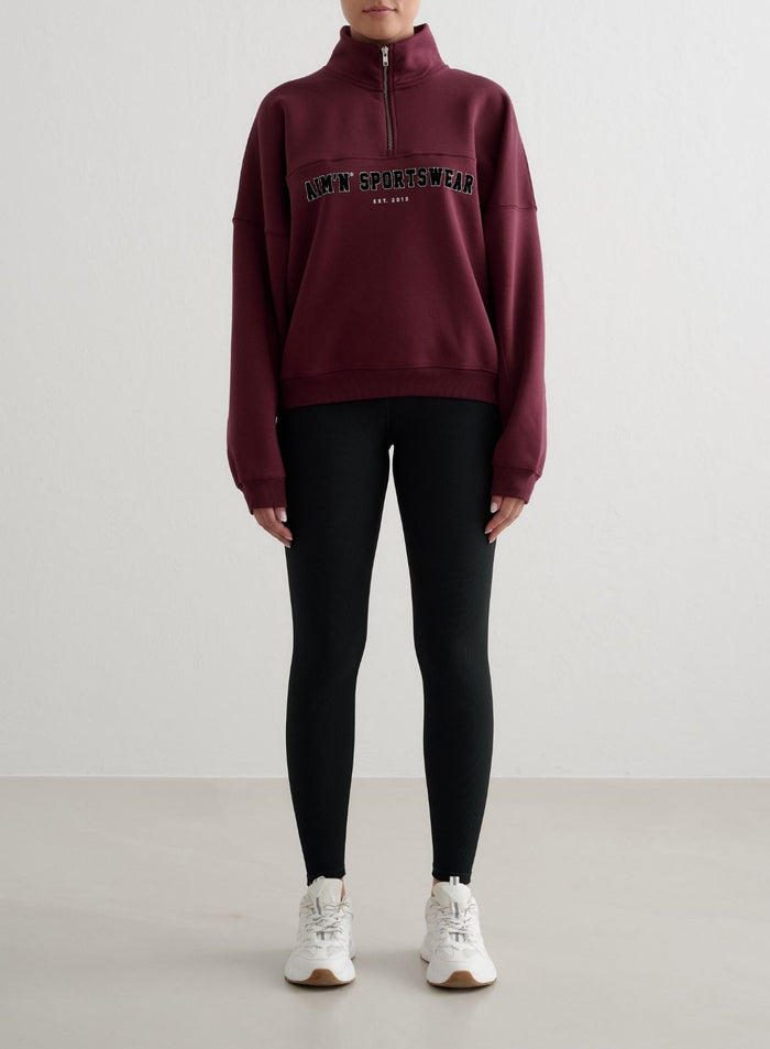 Cherry red aim'n Varsity Sweat Half Zip featuring a stand collar and varsity embroidery, paired with black leggings.