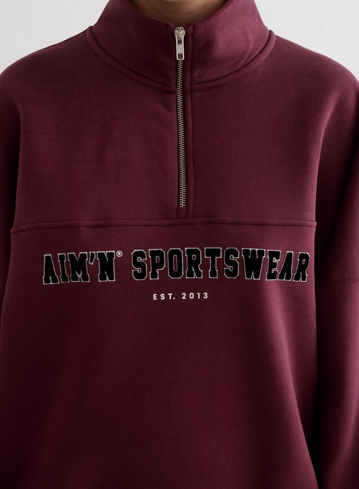Cherry red Varsity Sweat Half Zip with a stand collar and black varsity-style aim'n chest embroidery.