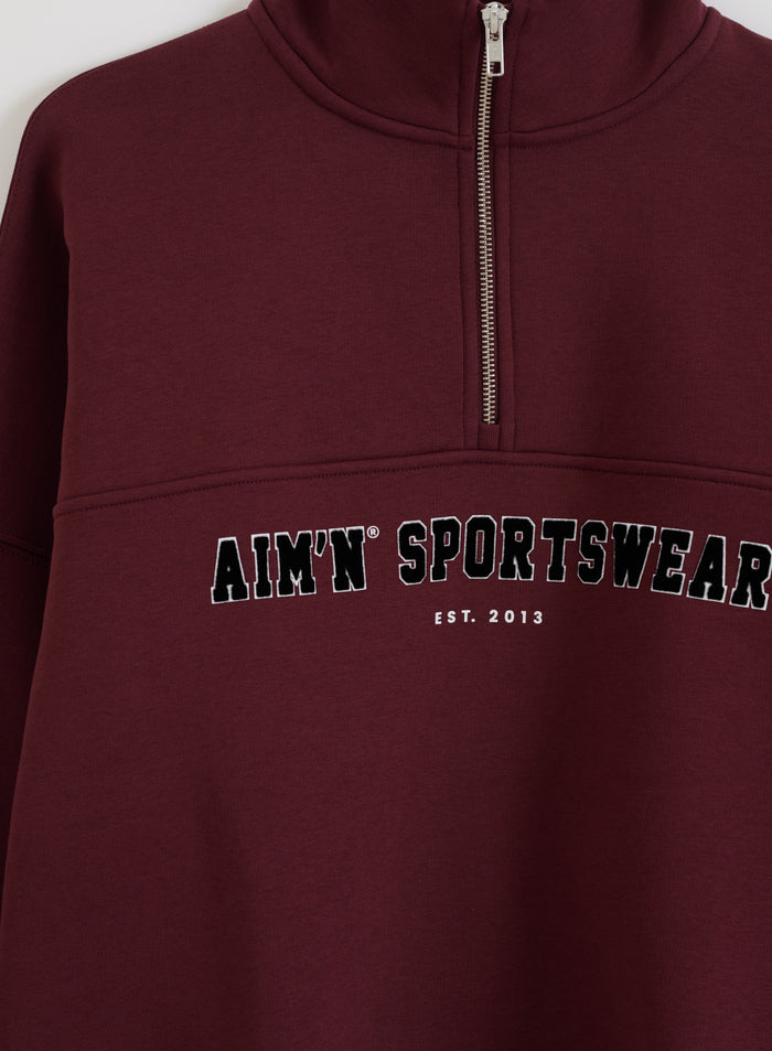 Cherry red varsity sweat half zip featuring a stand collar and aim'n sportswear chest print.