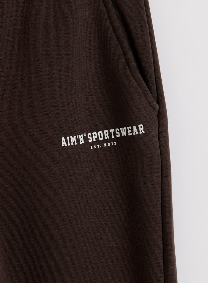 Close-up of chocolate brown Classic Sweatpants showing a side pocket and white aim'n logo on soft jersey.