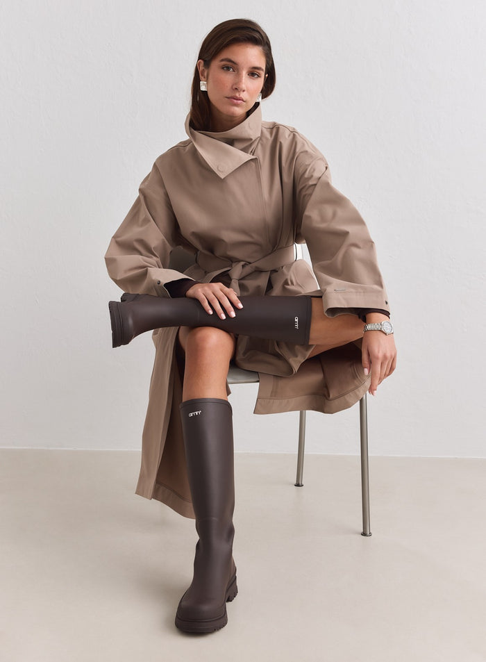 Knee-high chocolate rain boots by aim'n with a matte finish, paired with a stylish beige trench coat.