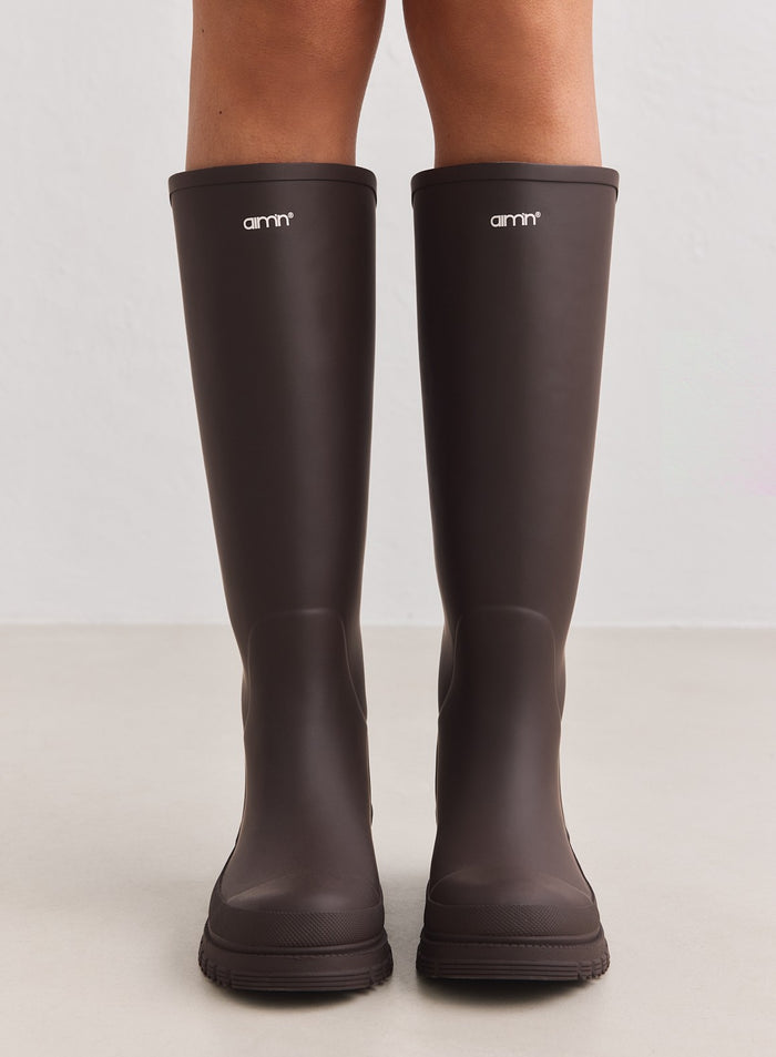 Knee-high chocolate matte rubber rain boots featuring the aim'n logo and reinforced toes.