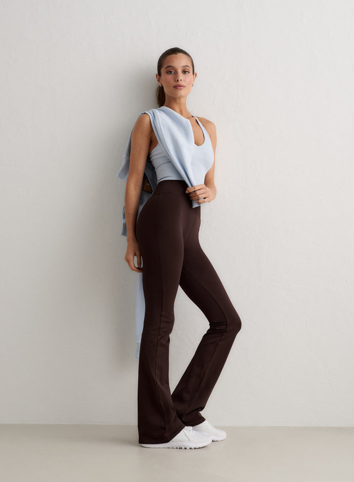 Woman posing against a white wall wearing chocolate brown aim'n Shape Seamless Flare Tall Tights, a light blue top and white sneakers [S-174cm]