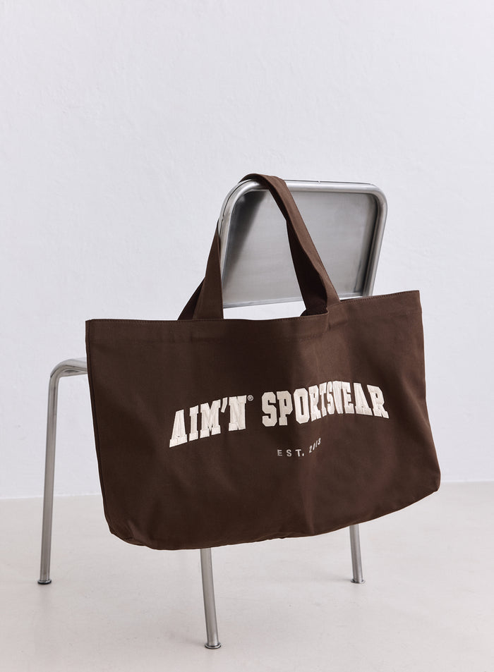 Chocolate brown Varsity Tote Bag in cotton canvas with a cream aim'n logo on a metal chair. [ONE-SIZE-165CM]
