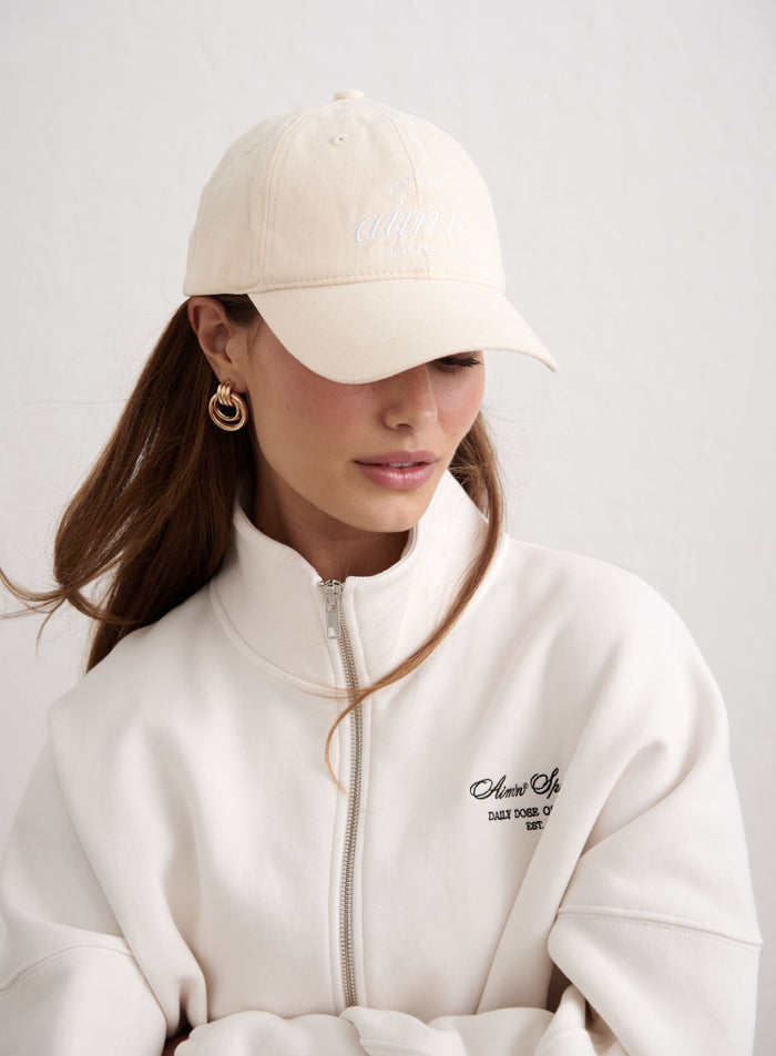 Sand-colored aim'n Club Cap with white embroidery, styled with a white zip-up sweatshirt.