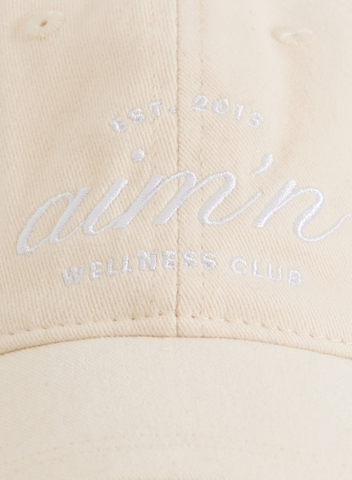 Sand cotton Club Cap with a white embroidered aim'n logo resting on a silver metal chair.