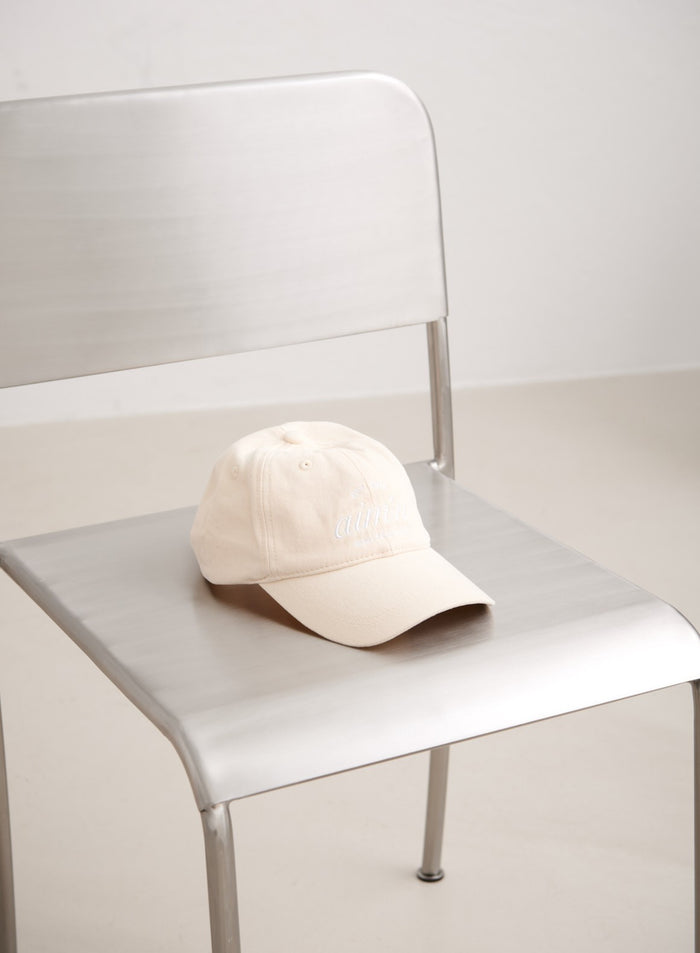 Sand-colored cotton Club Cap with white embroidered aim'n logo resting on a minimalist silver chair.