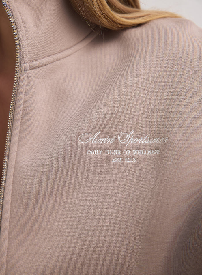 Close-up of the white aim'n script logo on a dark clay zip jacket made of soft cotton blend. [M-170cm]
