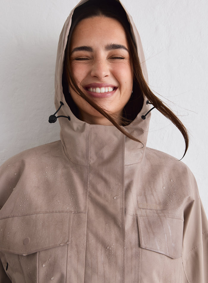 A woman smiling while wearing a Dark Clay Waterproof Jacket with the hood up and water droplets on the fabric [M-174cm]