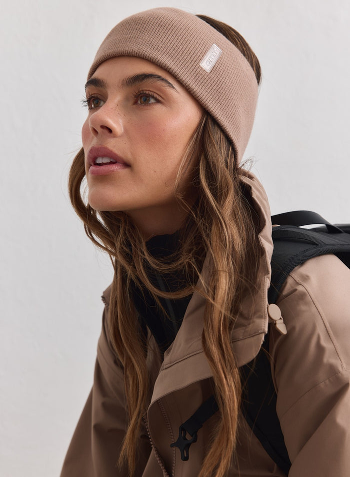 Hazelnut ribbed knit Outdoor Logo Headband with white aim'n branding, worn with a beige jacket and backpack.