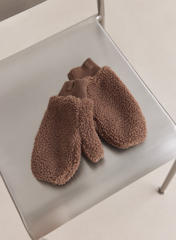Hazelnut pile mittens from aim'n featuring a fuzzy texture and ribbed cuffs, resting on a metal chair.