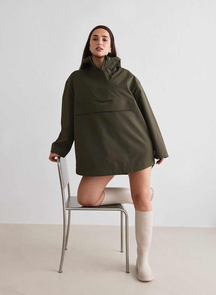 Model wearing a Khaki Waterproof Oversized Anorak by aim'n and tall beige boots, posing with one knee on a chair [M-174cm]