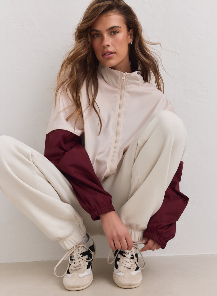 Merlot and beige aim'n Balance Windstopper Tracksuit Jacket worn by a woman crouching against a white wall.