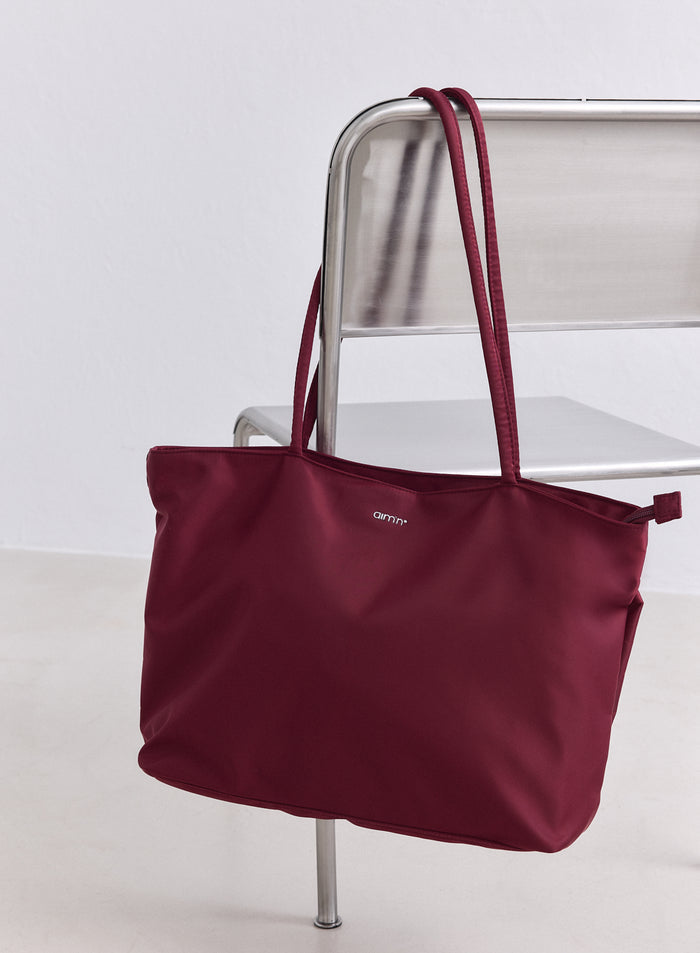 Merlot nylon tote bag by aim'n hanging on a silver chair in a minimalist room.