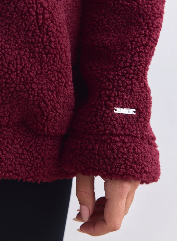 Close-up of the merlot teddy pile fabric and a small silver aim'n logo plate on the jacket cuff. [S-165cm]