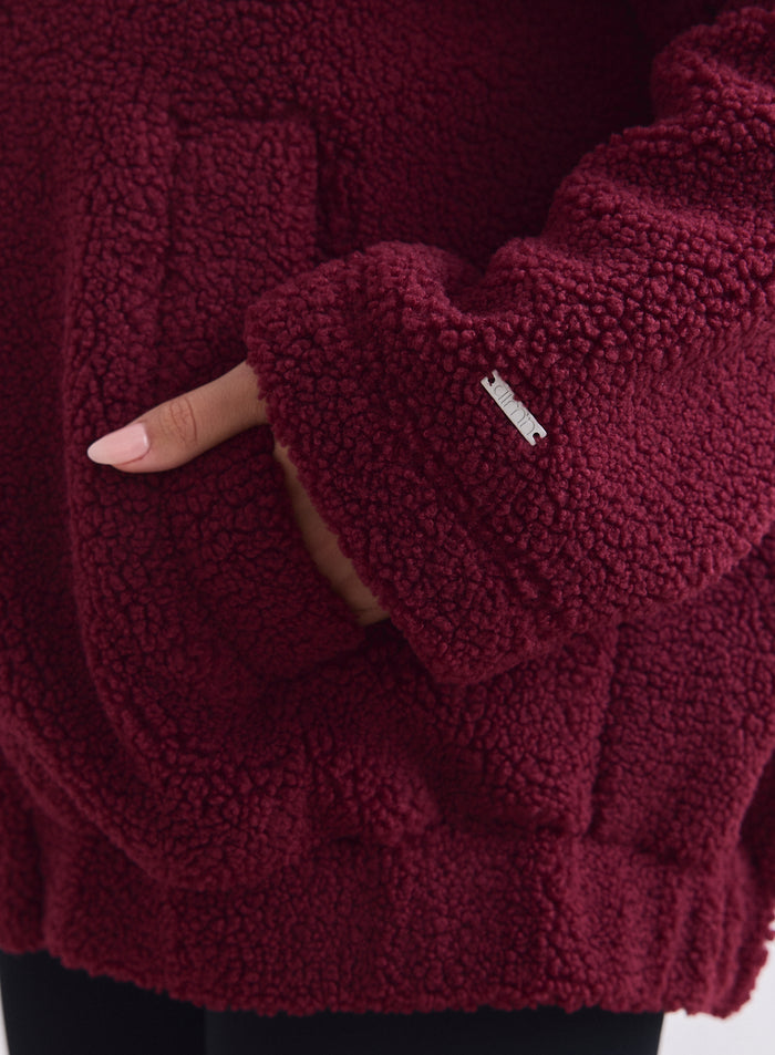 Close-up of a hand in the pocket of a merlot teddy pile jacket with a silver aim'n logo. [S-165cm]