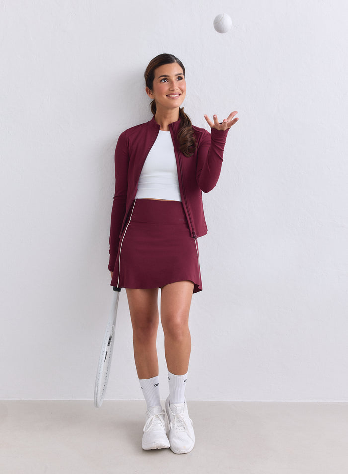 Woman wearing a merlot aim'n Sense Zip Jacket and matching skirt tossing a tennis ball. [XS-165cm]