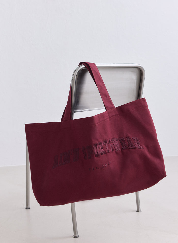 Merlot cotton varsity tote bag by aim'n featuring varsity branding hanging on a metal chair.