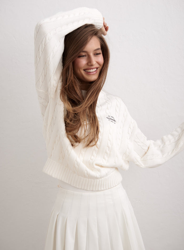 Off-white cotton cable-knit sweater by aim'n styled with a white pleated skirt on a smiling model. [M-174cm]