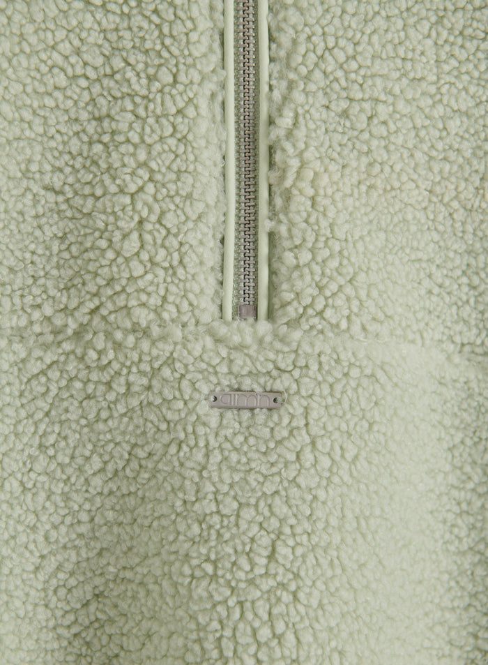Close-up detail of the pistachio green fleece fabric, zipper, and aim'n logo plate on the Pile Half Zip